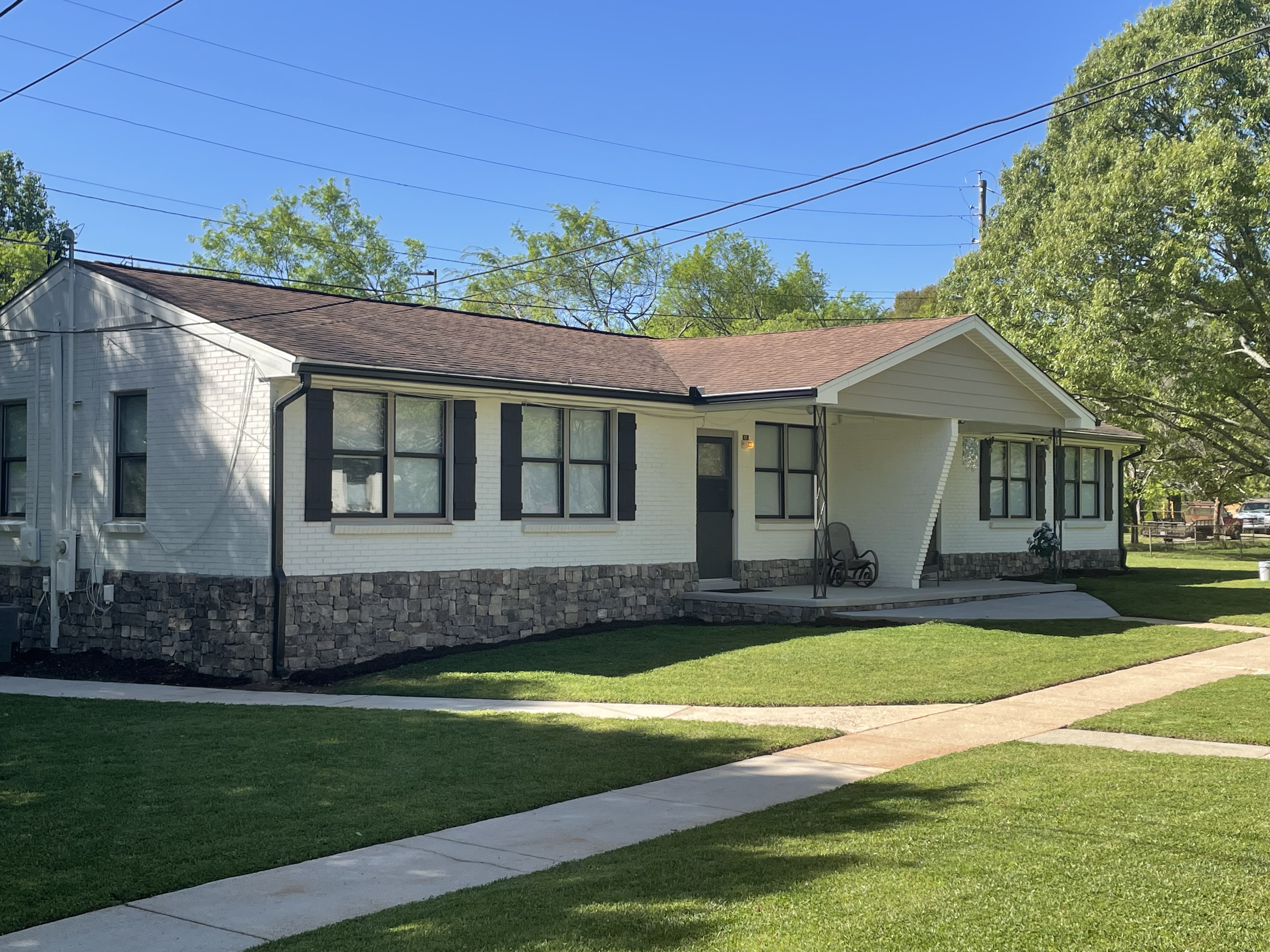 SHows the front yard of a multiple entrance brick row house. Also shows several trees and bushes in the yard